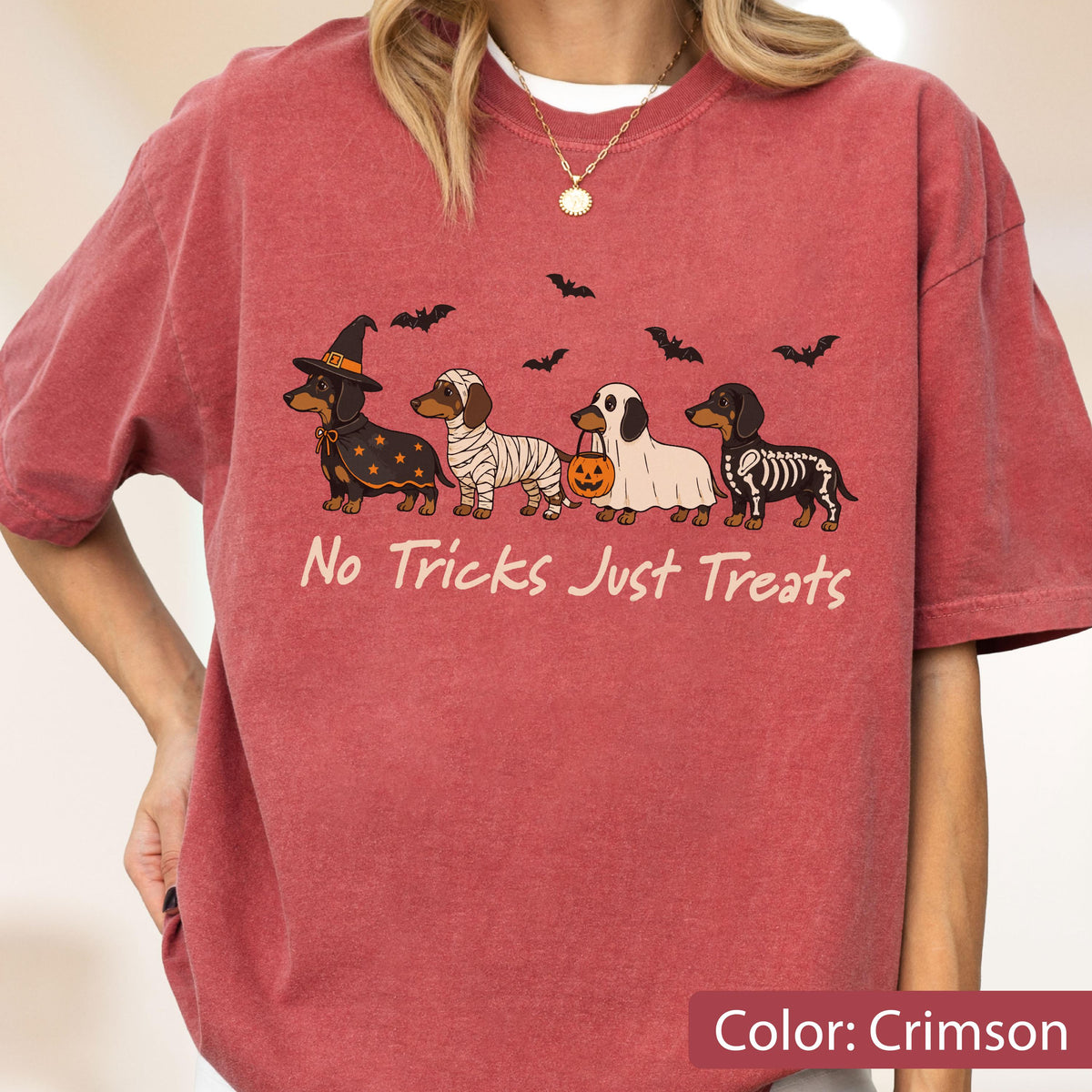 May include: A crimson short-sleeve t-shirt featuring a graphic of four dachshunds in Halloween costumes, including a witch, mummy, ghost, and skeleton. Above the dogs are black bats, and below is the text No Tricks Just Treats in cream.