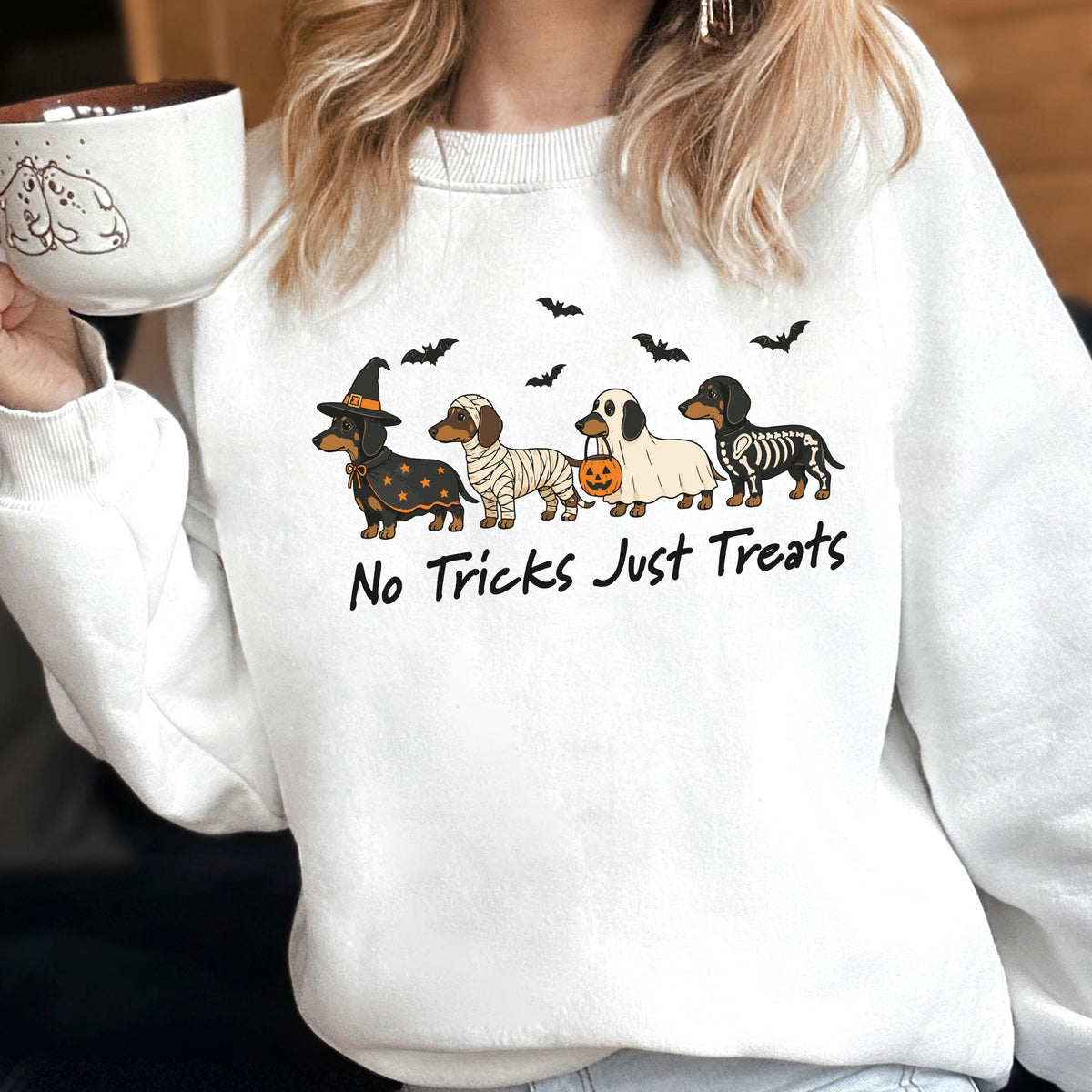 May include: A white crewneck sweatshirt featuring a graphic of four dachshunds in Halloween costumes: a witch, a mummy, a ghost holding a pumpkin bucket, and a skeleton. Above the dogs are black bats, and below is the text No Tricks Just Treats.