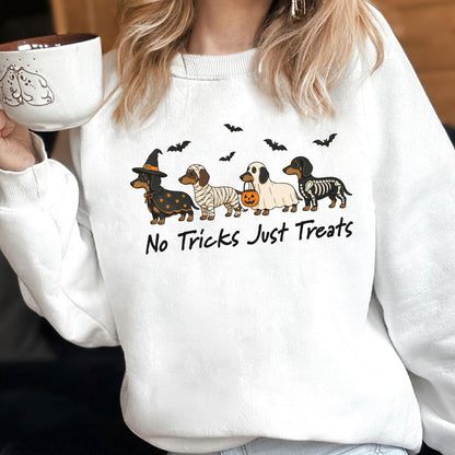 May include: A white crewneck sweatshirt featuring a graphic of four dachshunds in Halloween costumes: a witch, a mummy, a ghost holding a pumpkin bucket, and a skeleton. Above the dogs are black bats, and below is the text No Tricks Just Treats.
