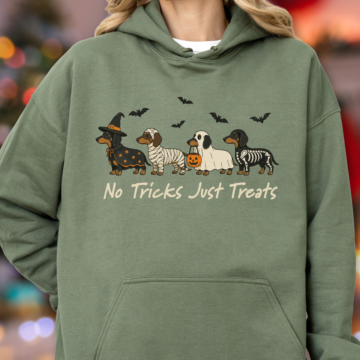 May include: An olive green hooded sweatshirt featuring a graphic of four dachshunds in Halloween costumes, including a witch, a mummy, a ghost holding a pumpkin bucket, and a skeleton. The text "No Tricks Just Treats" is printed below the dogs.