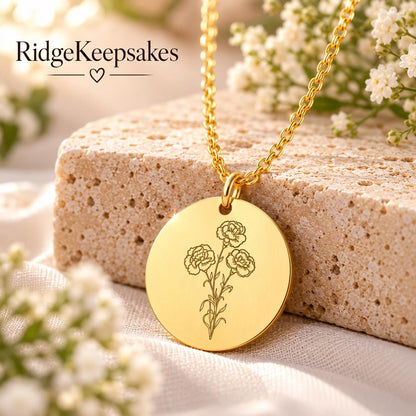 Close-up of a round gold-tone pendant engraved with a delicate flower stem, hanging from a matching chain against a neutral stone and floral background.