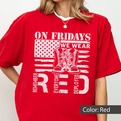 May include: A bright red short-sleeved t-shirt featuring a white graphic print of a stylized American flag, a pair of military boots, and the text ON FRIDAYS WE WEAR RED REMEMBER EVERYONE DEPLOYED. The design is centered on the front of the shirt.