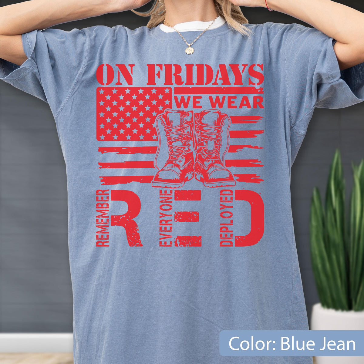 May include: A blue jean colored t-shirt featuring a red graphic print. The design includes a stylized American flag, a pair of military boots, and the text ON FRIDAYS WE WEAR RED REMEMBER EVERYONE DEPLOYED in a distressed, bold red font.