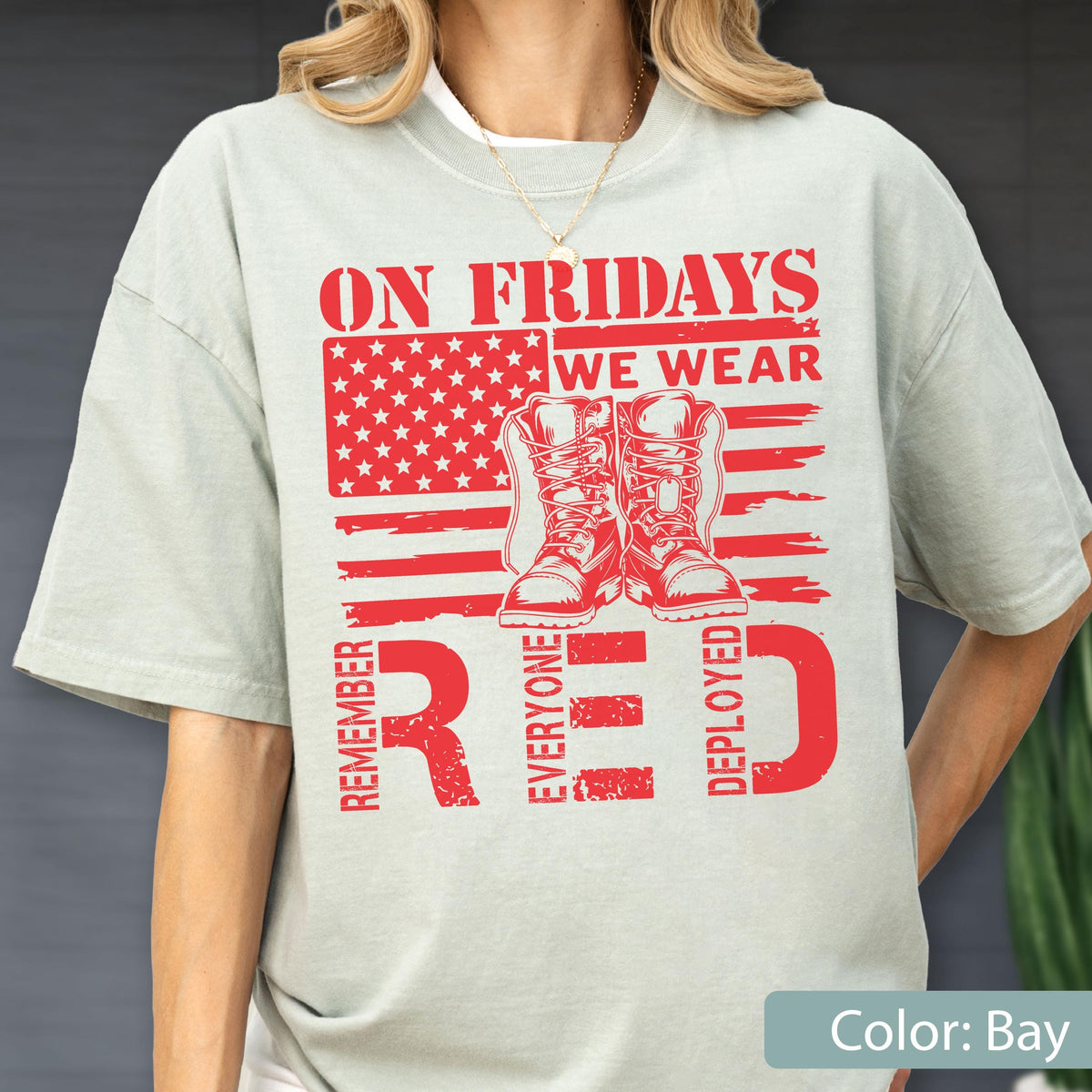 May include: A light sage green t-shirt featuring a red graphic print of a stylized American flag with a pair of combat boots in the center. The text reads ON FRIDAYS WE WEAR RED and REMEMBER EVERYONE DEPLOYED in bold, distressed red lettering.