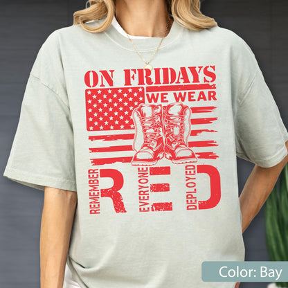 May include: A light sage green t-shirt featuring a red graphic print of a stylized American flag with a pair of combat boots in the center. The text reads ON FRIDAYS WE WEAR RED and REMEMBER EVERYONE DEPLOYED in bold, distressed red lettering.