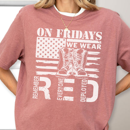 May include: A dusty rose short-sleeve t-shirt featuring a white graphic print of an American flag, a pair of military combat boots, and the text ON FRIDAYS WE WEAR RED REMEMBER EVERYONE DEPLOYED in a distressed, bold, block-style font.