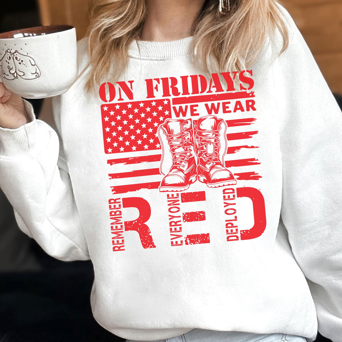 May include: A white crewneck sweatshirt featuring a red graphic print. The design includes a stylized American flag, a pair of military combat boots, and the text: ON FRIDAYS WE WEAR RED REMEMBER EVERYONE DEPLOYED in a bold, distressed red font.