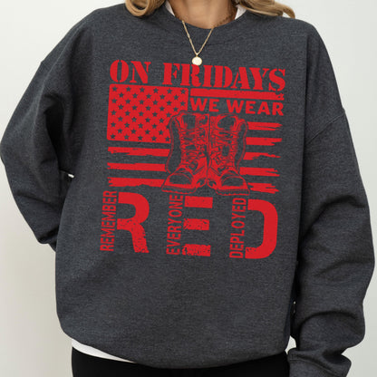 May include: A dark charcoal gray crewneck sweatshirt featuring a red graphic print. The design includes a stylized American flag, a pair of combat boots, and the text: ON FRIDAYS WE WEAR RED, REMEMBER EVERYONE DEPLOYED, arranged in a bold layout.