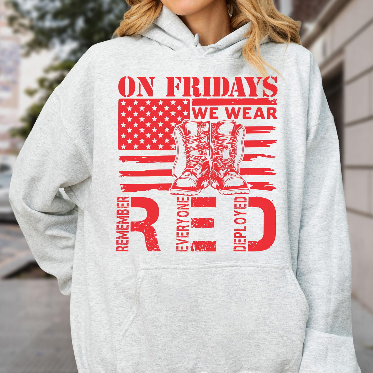 May include: A light gray hooded sweatshirt featuring a red graphic print. The design includes a stylized American flag, a pair of military boots, and the text: ON FRIDAYS WE WEAR RED, REMEMBER EVERYONE DEPLOYED, arranged in a bold, distressed font.