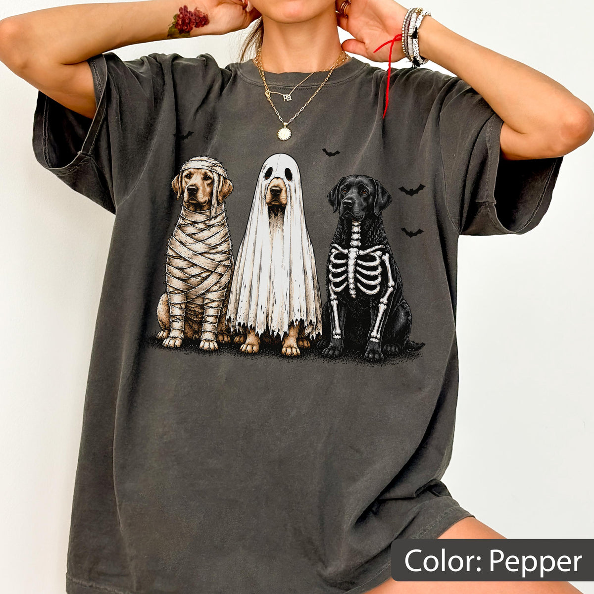 May include: A pepper-colored t-shirt featuring a graphic of three dogs in Halloween costumes: one wrapped like a mummy, one wearing a white ghost sheet, and one with a skeleton print on its chest, surrounded by small black bat silhouettes.