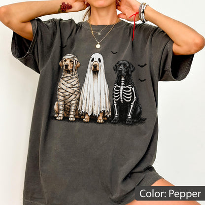 May include: A pepper-colored t-shirt featuring a graphic of three dogs in Halloween costumes: one wrapped like a mummy, one wearing a white ghost sheet, and one with a skeleton print on its chest, surrounded by small black bat silhouettes.