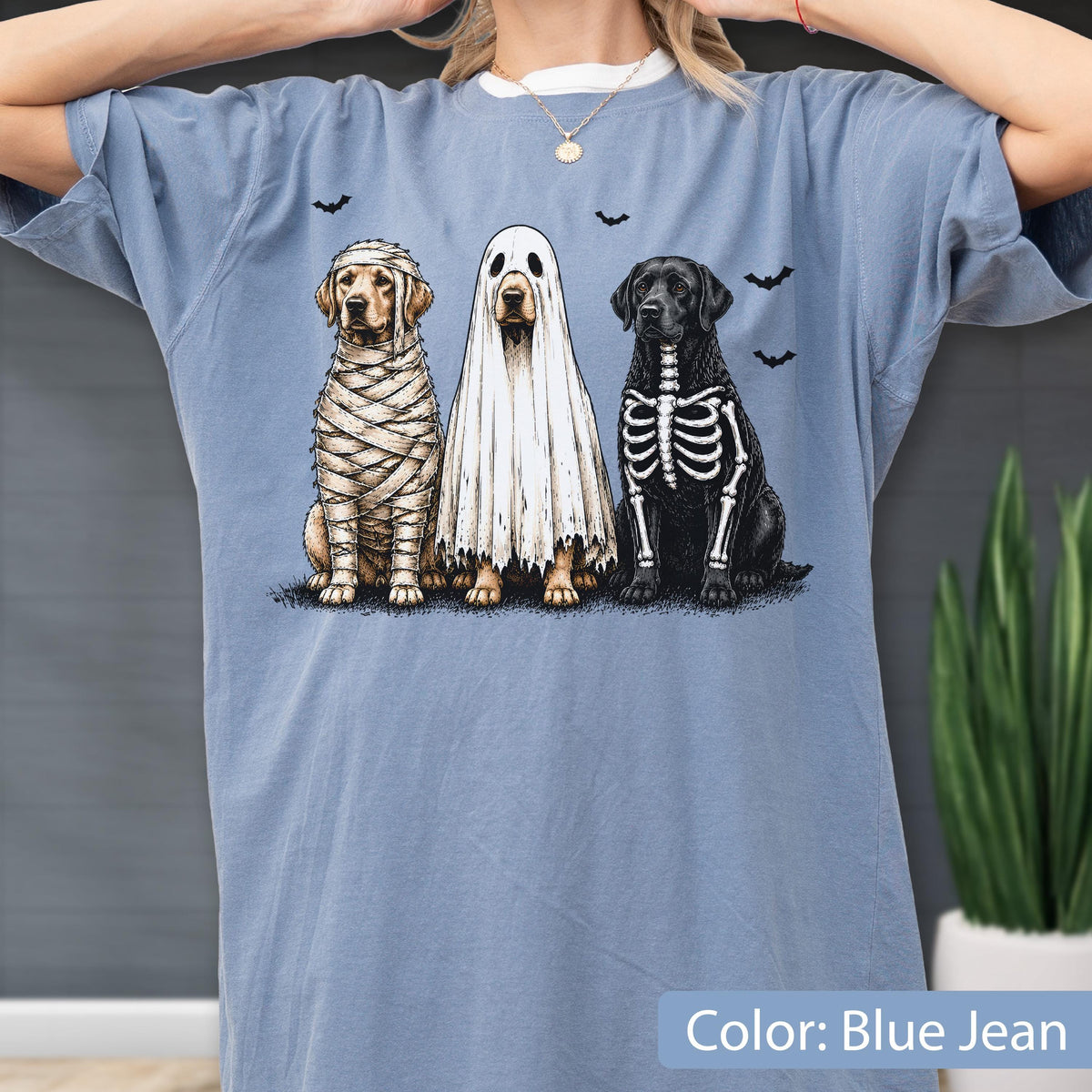 May include: A blue jean colored t-shirt featuring a graphic of three dogs dressed for Halloween. One dog is wrapped as a mummy, one is wearing a white ghost sheet, and one has a skeleton print on its black fur, surrounded by small black bats.