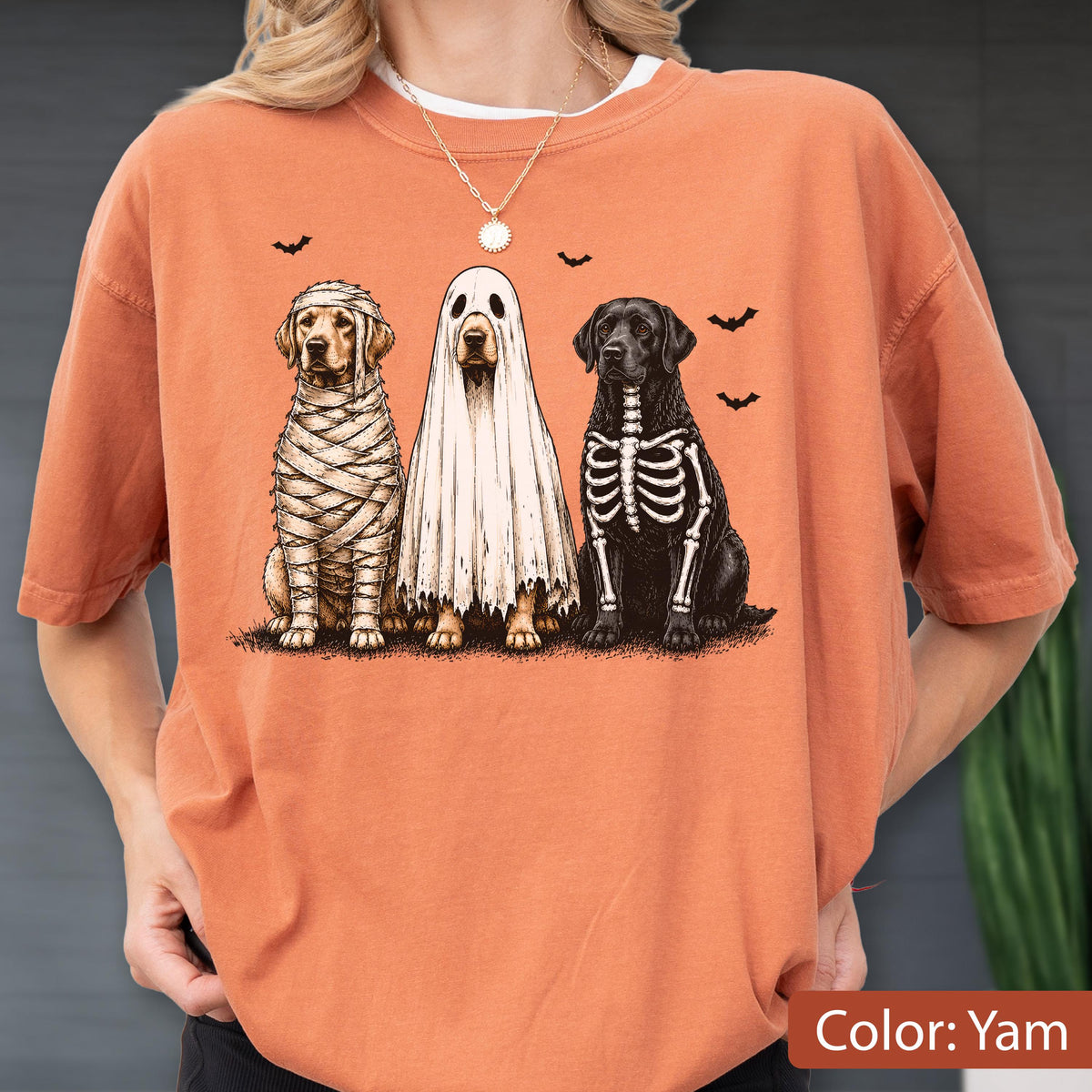 May include: An orange t-shirt featuring a graphic of three dogs in Halloween costumes. One dog is wrapped like a mummy, one is wearing a white ghost sheet, and one has a skeleton print on its black fur. Small black bats are flying above the dogs.