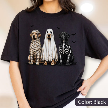 May include: A black t-shirt featuring a graphic of three dogs in Halloween costumes. One dog is wrapped in mummy bandages, one is wearing a white ghost sheet, and one has a white skeleton print on its black fur, with small black bats nearby.