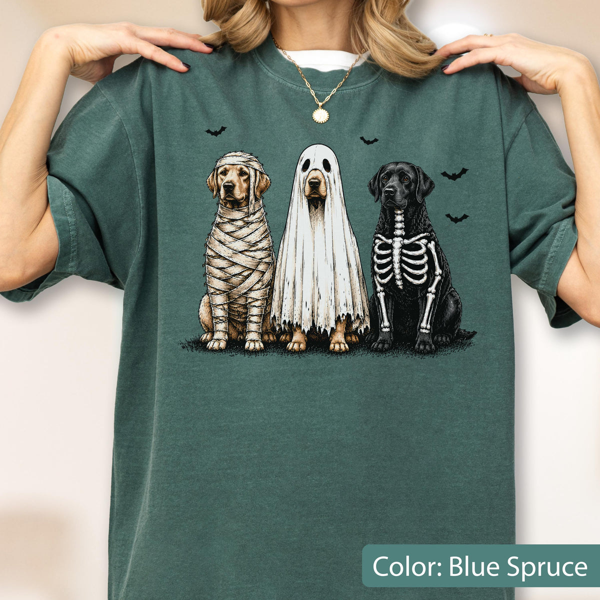 May include: A blue spruce crewneck t-shirt featuring a graphic of three dogs in Halloween costumes: one wrapped like a mummy, one wearing a white ghost sheet, and one with a skeleton print, surrounded by small black bat silhouettes.