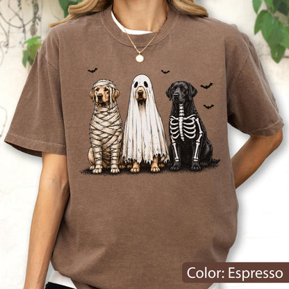 May include: An espresso-colored t-shirt featuring a graphic of three dogs in Halloween costumes. One dog is wrapped like a mummy, one is wearing a white ghost sheet, and one has a skeleton print on its black fur. Small black bats fly above.