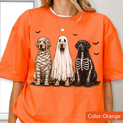 May include: An orange t-shirt featuring a graphic print of three dogs in Halloween costumes. One dog is wrapped like a mummy, one is wearing a white ghost sheet, and one has a skeleton print on its body. Small black bats are scattered above the dogs.