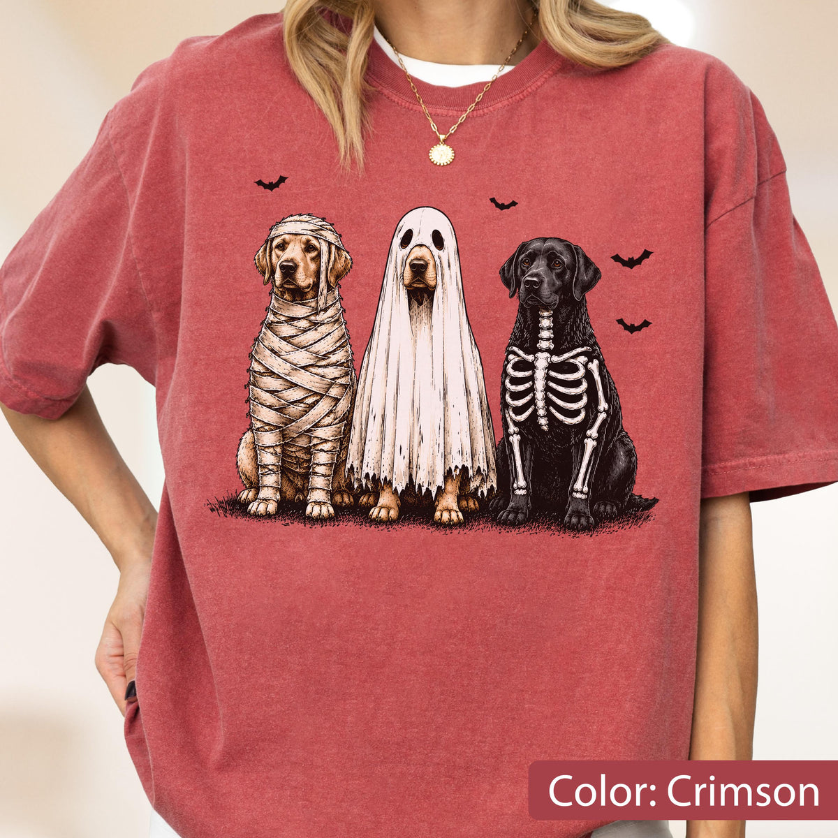 May include: A crimson t-shirt featuring a graphic of three dogs dressed for Halloween. One dog is wrapped in mummy bandages, one is wearing a white ghost sheet, and one has a white skeleton print on its black fur, with small black bats above.