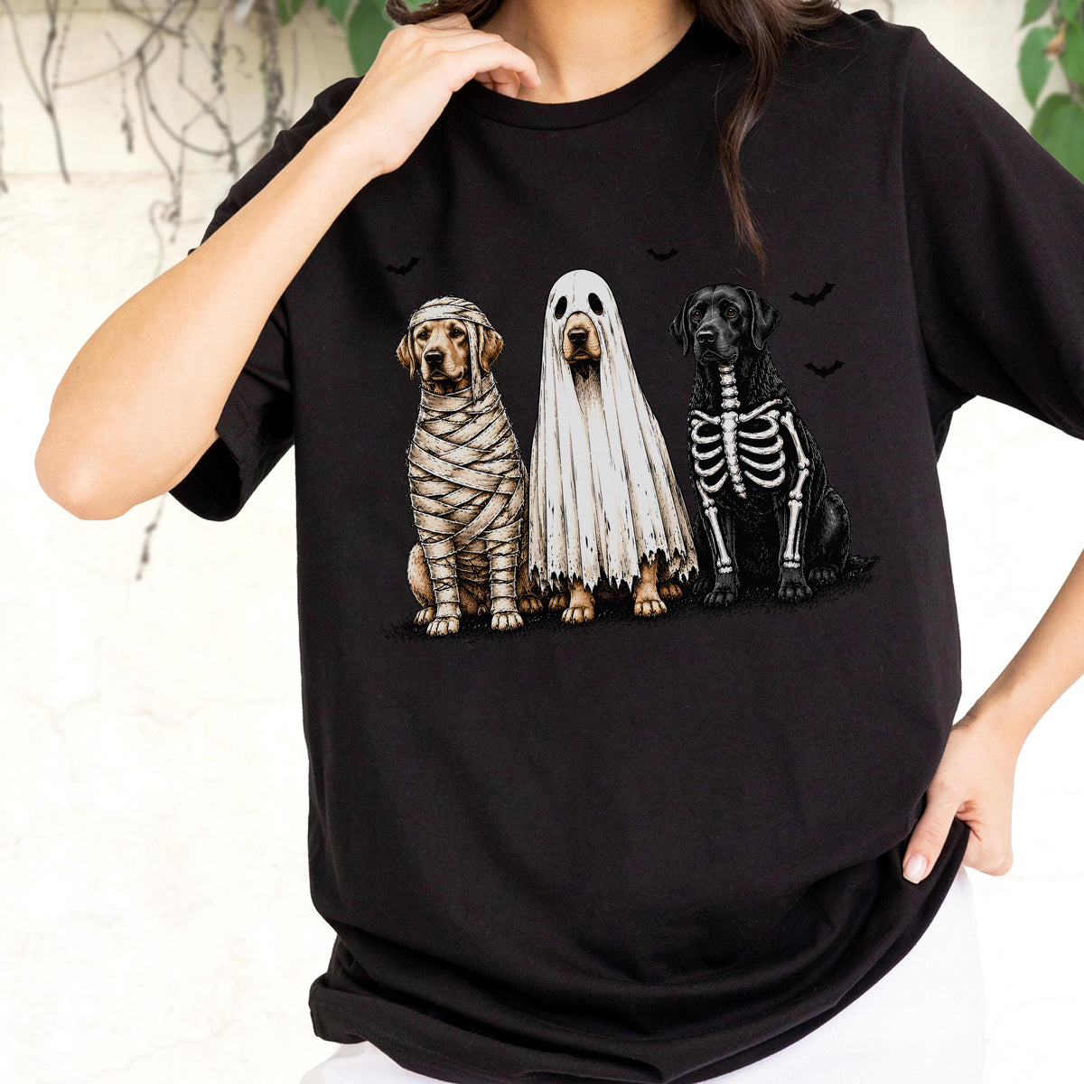 May include: A black short-sleeved t-shirt featuring a graphic of three dogs in Halloween costumes. One dog is wrapped in mummy bandages, the middle dog is wearing a white ghost sheet, and the third dog has a white skeleton print on its black fur.