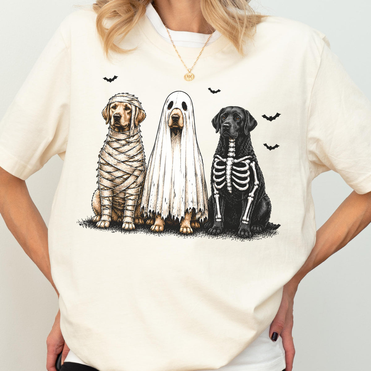 May include: A cream-colored t-shirt featuring a graphic of three dogs in Halloween costumes. One dog is wrapped like a mummy, one is wearing a white ghost sheet, and one has a skeleton print on its chest. Small black bats are scattered above.