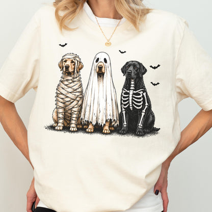 May include: A cream-colored t-shirt featuring a graphic of three dogs in Halloween costumes. One dog is wrapped like a mummy, one is wearing a white ghost sheet, and one has a skeleton print on its chest. Small black bats are scattered above.