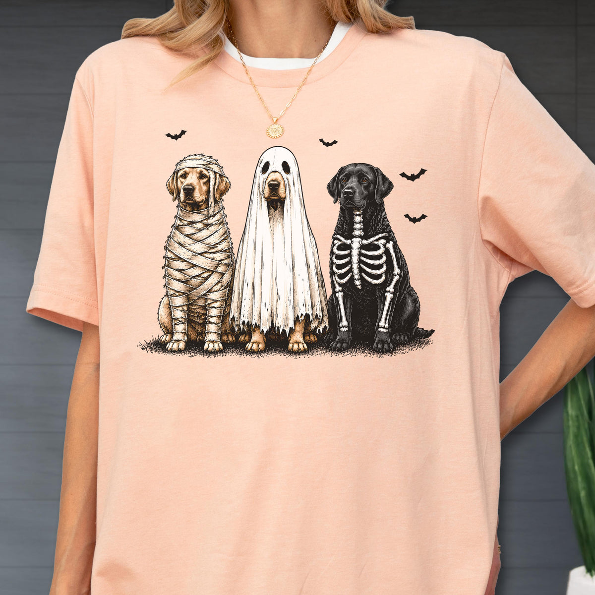 May include: A peach-colored t-shirt featuring a graphic of three dogs in Halloween costumes: one wrapped like a mummy, one wearing a white ghost sheet, and one with a skeleton print. Small black bats are scattered above the dogs.