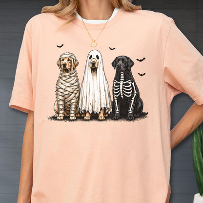 May include: A peach-colored t-shirt featuring a graphic of three dogs in Halloween costumes: one wrapped like a mummy, one wearing a white ghost sheet, and one with a skeleton print. Small black bats are scattered above the dogs.