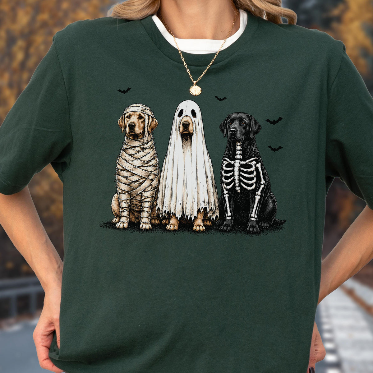May include: A dark green t-shirt featuring a graphic of three dogs in Halloween costumes. One dog is wrapped like a mummy, one is wearing a white ghost sheet, and one has a skeleton print on its black fur. Small black bats are printed above them.