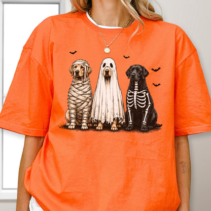 May include: A bright orange short-sleeved t-shirt featuring a graphic of three dogs dressed for Halloween. One dog is wrapped like a mummy, one is wearing a white ghost sheet, and the third has a skeleton print on its black fur, with small bats.