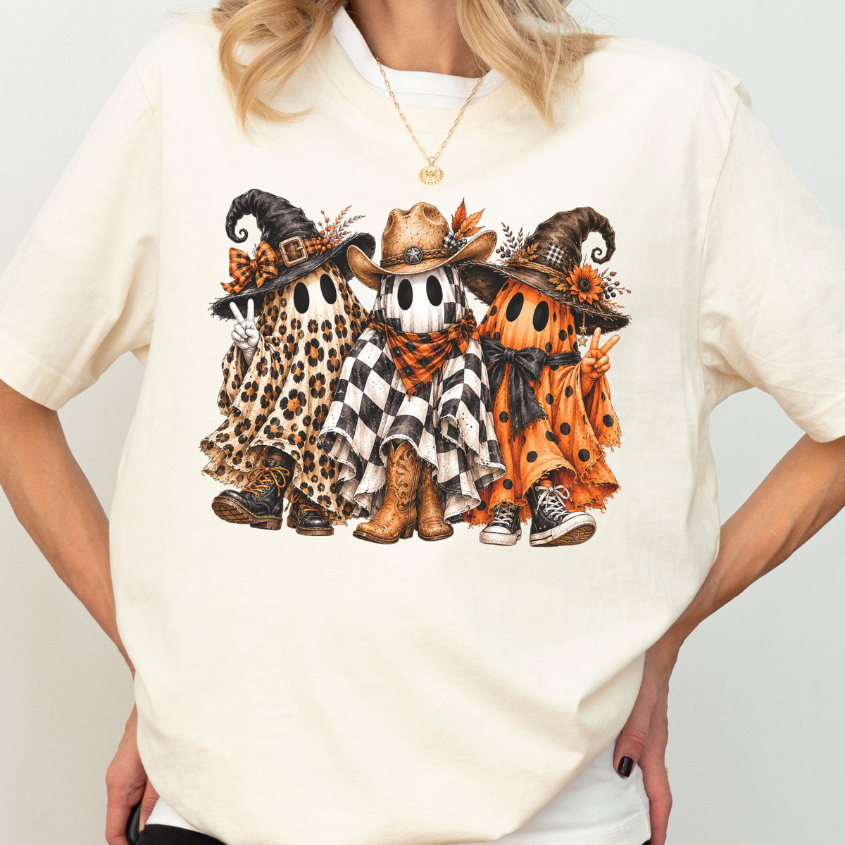 May include: A cream-colored short-sleeve t-shirt featuring a graphic of three ghosts wearing hats. The left ghost has a leopard print sheet and boots, the center ghost wears a cowboy hat and checkered sheet, and the right ghost has an orange polka dot sheet.