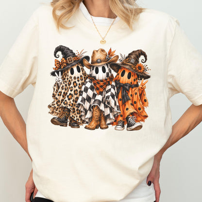 May include: A cream-colored short-sleeve t-shirt featuring a graphic of three ghosts wearing hats. The left ghost has a leopard print sheet and boots, the center ghost wears a cowboy hat and checkered sheet, and the right ghost has an orange polka dot sheet.