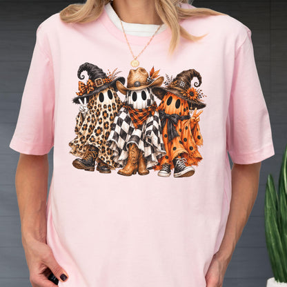 May include: A light pink t-shirt featuring a graphic of three ghosts wearing hats. The left ghost has a leopard print sheet and boots, the center ghost has a checkered sheet and cowboy boots, and the right ghost has an orange polka dot sheet.