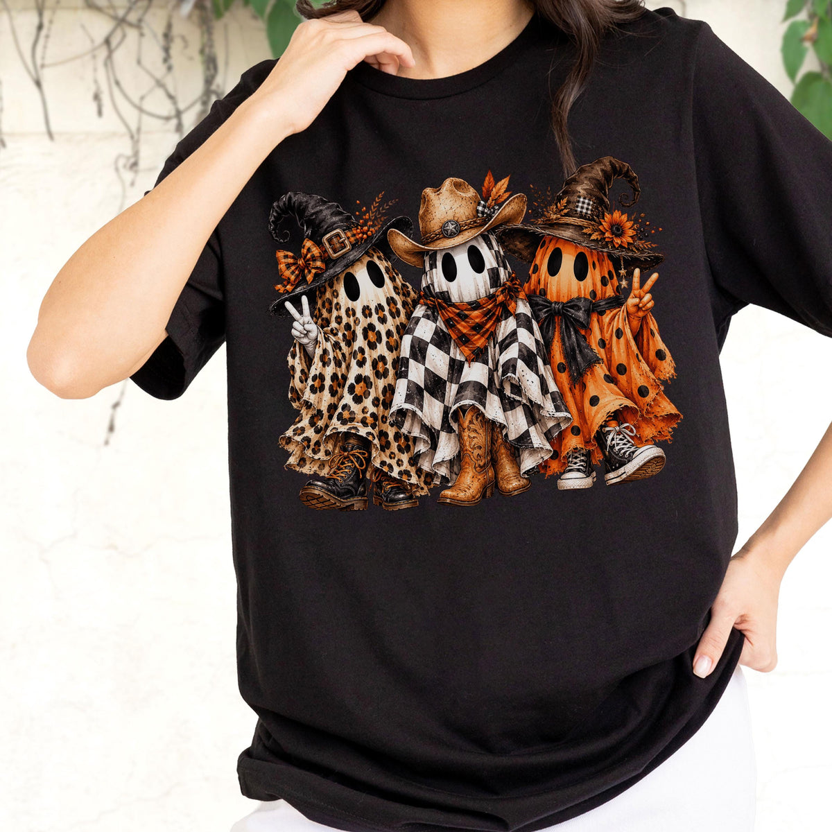 May include: A black t-shirt featuring a graphic of three ghosts wearing hats and footwear. The left ghost has a leopard print sheet, the middle ghost has a checkered sheet and cowboy hat, and the right ghost has an orange polka dot sheet.