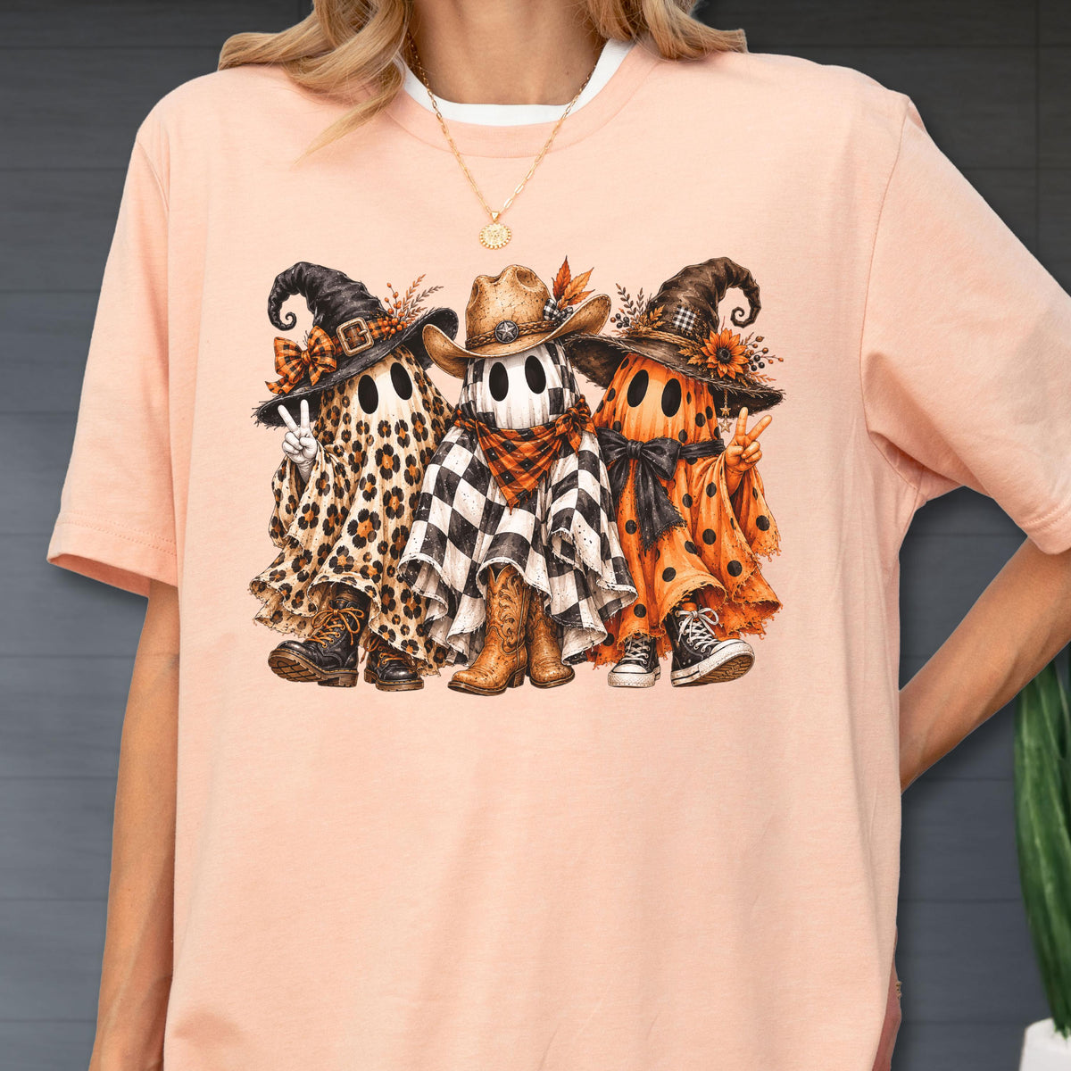 May include: A peach-colored t-shirt featuring a graphic of three ghosts wearing hats and footwear. The left ghost has a leopard print sheet, the middle ghost has a black and white checkered sheet with a cowboy hat, and the right ghost has an orange polka dot sheet.