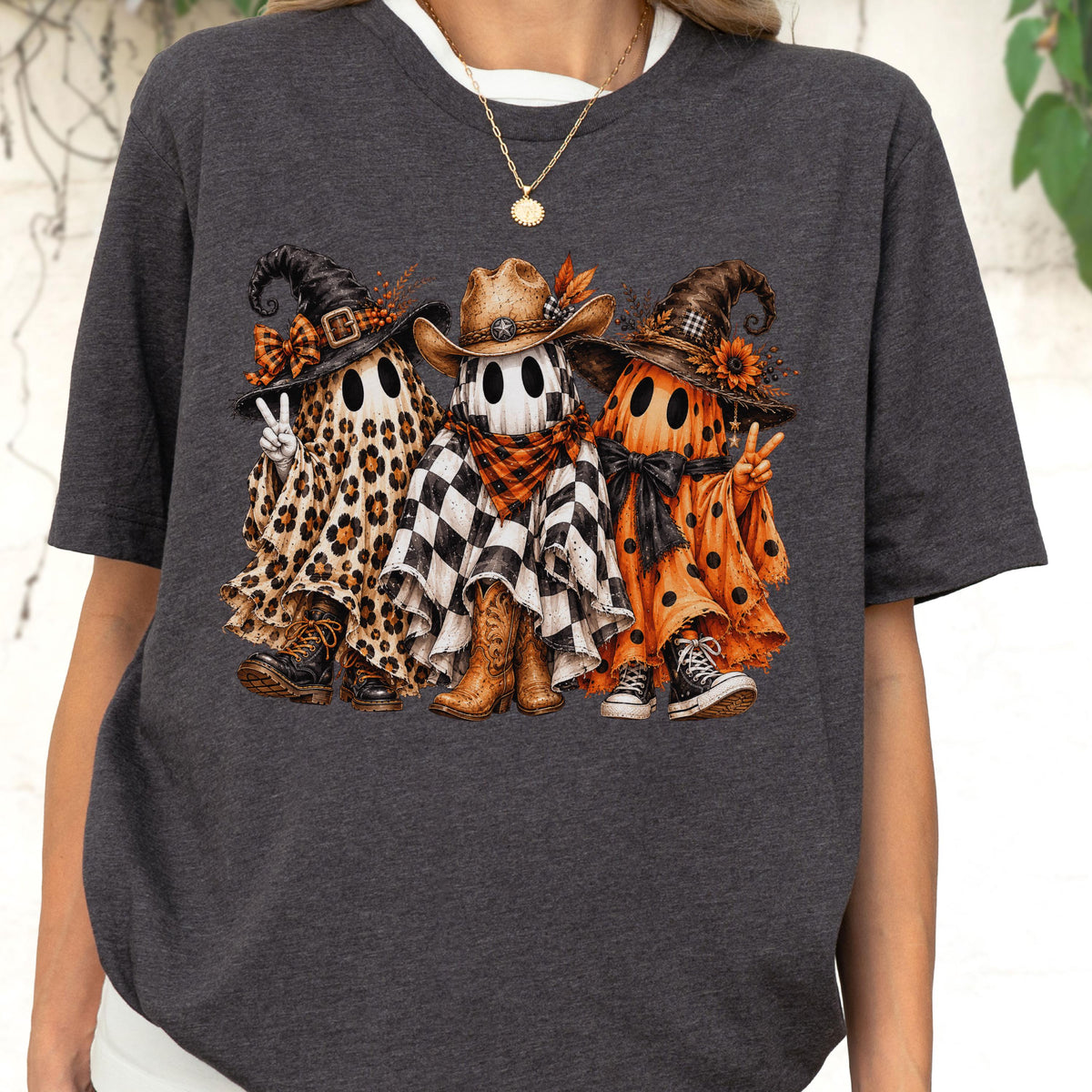 May include: A dark heather gray t-shirt featuring a graphic of three ghosts wearing hats and boots. The left ghost has a leopard print sheet, the middle ghost has a black and white buffalo check sheet, and the right ghost has an orange polka dot sheet.