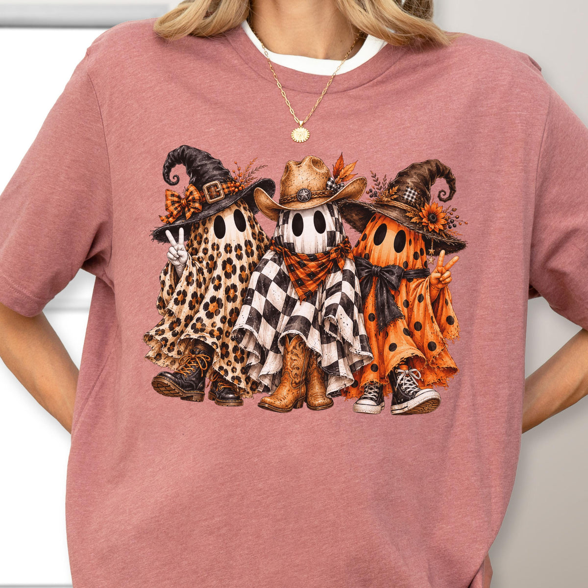 May include: A mauve short-sleeved t-shirt featuring a graphic of three ghosts. The left ghost wears a leopard print sheet and witch hat, the center ghost wears a checkered sheet and cowboy hat, and the right ghost wears an orange polka dot sheet.