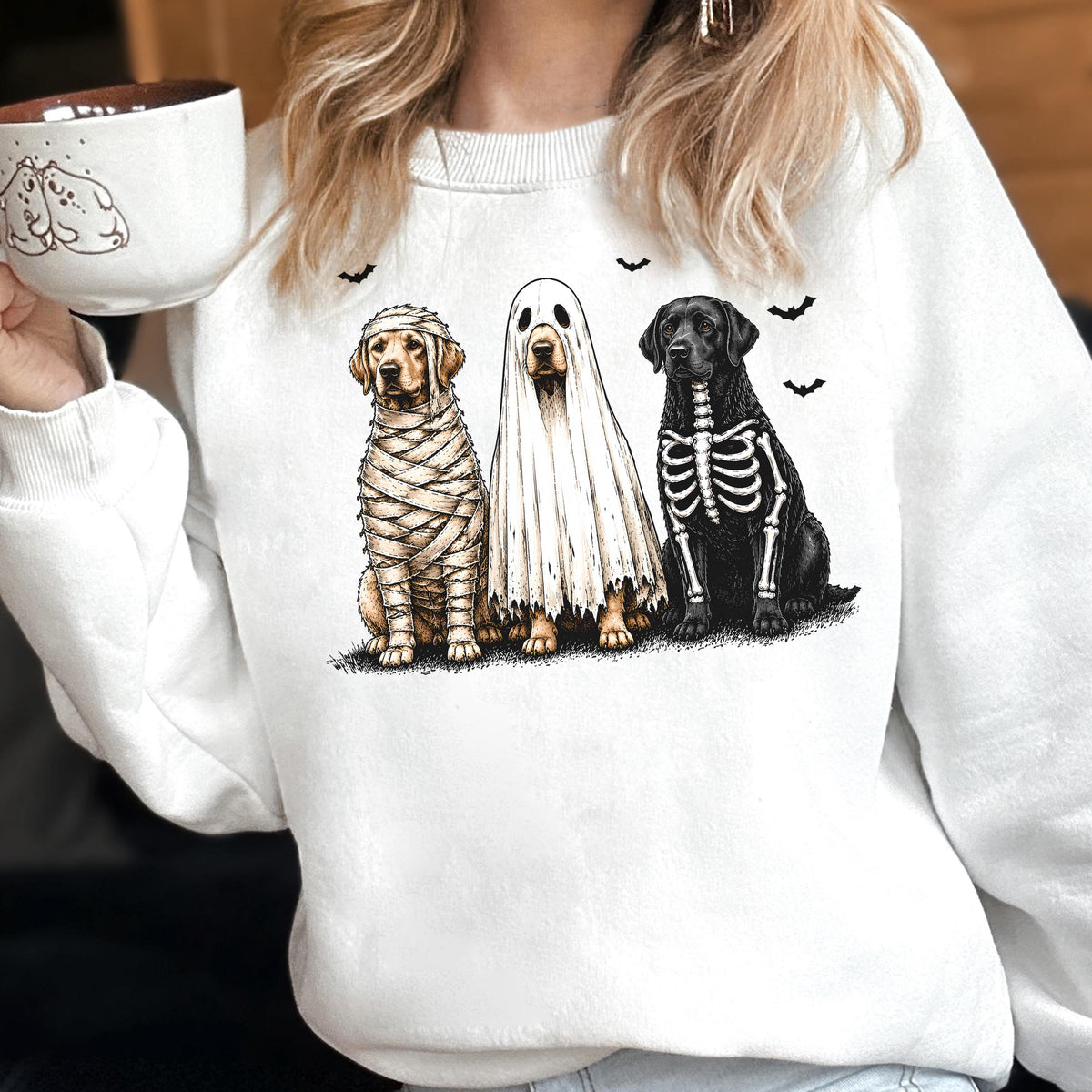 May include: A white crewneck sweatshirt featuring a graphic of three dogs in Halloween costumes: one wrapped as a mummy, one wearing a ghost sheet, and one with a skeleton print. Small black bats are scattered above the dogs.
