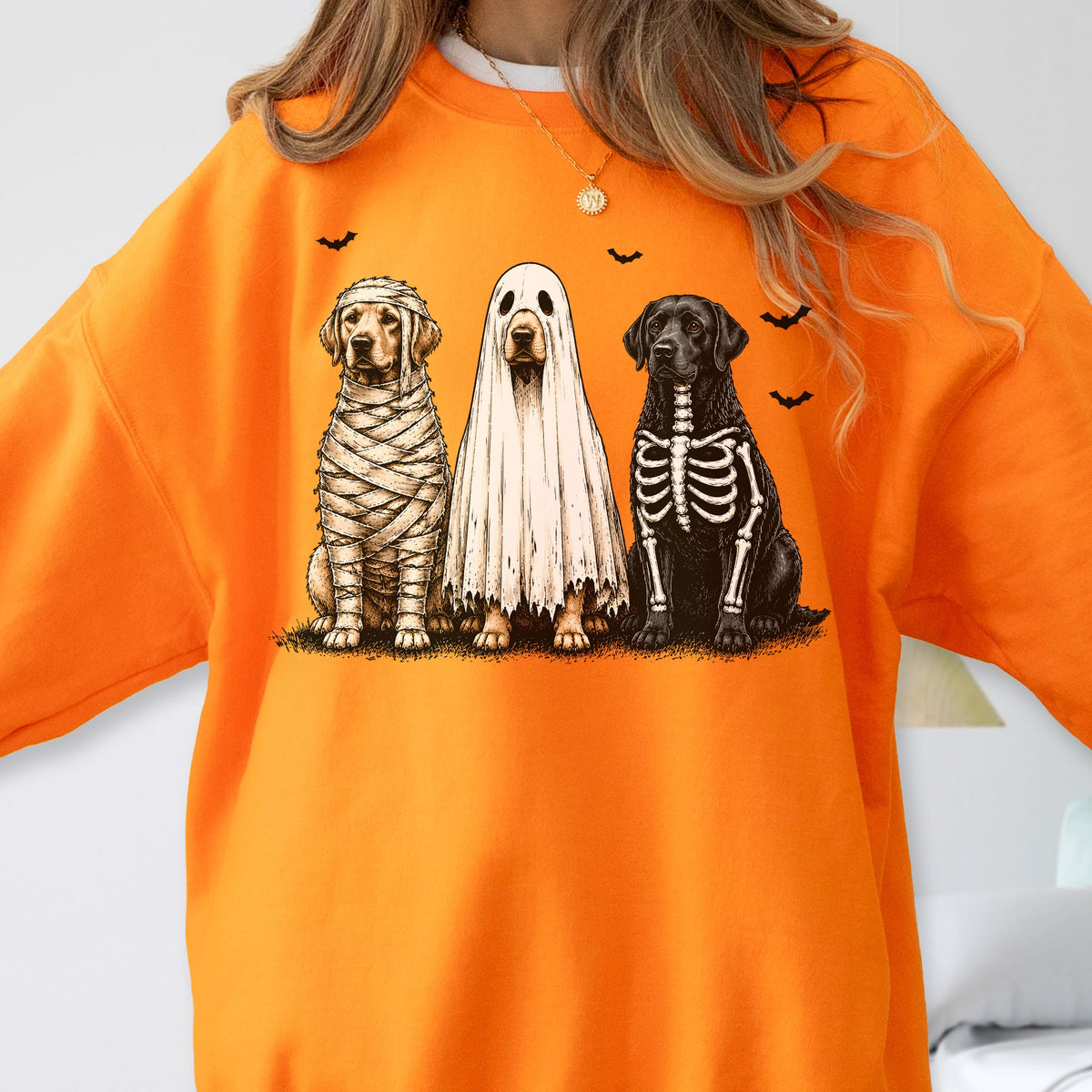 May include: A bright orange crewneck sweatshirt featuring a graphic print of three dogs in Halloween costumes. One dog is wrapped like a mummy, one is wearing a white ghost sheet, and one has a skeleton ribcage design. Small black bats fly above.