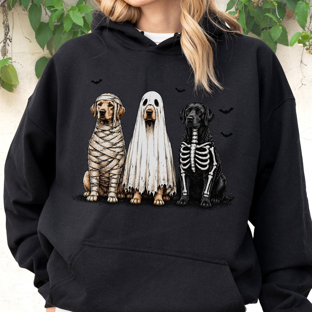 May include: A black hooded sweatshirt featuring a graphic print of three dogs dressed for Halloween. One dog is wrapped like a mummy, one is wearing a white ghost sheet, and one has a skeleton ribcage design. Small black bats are printed above.