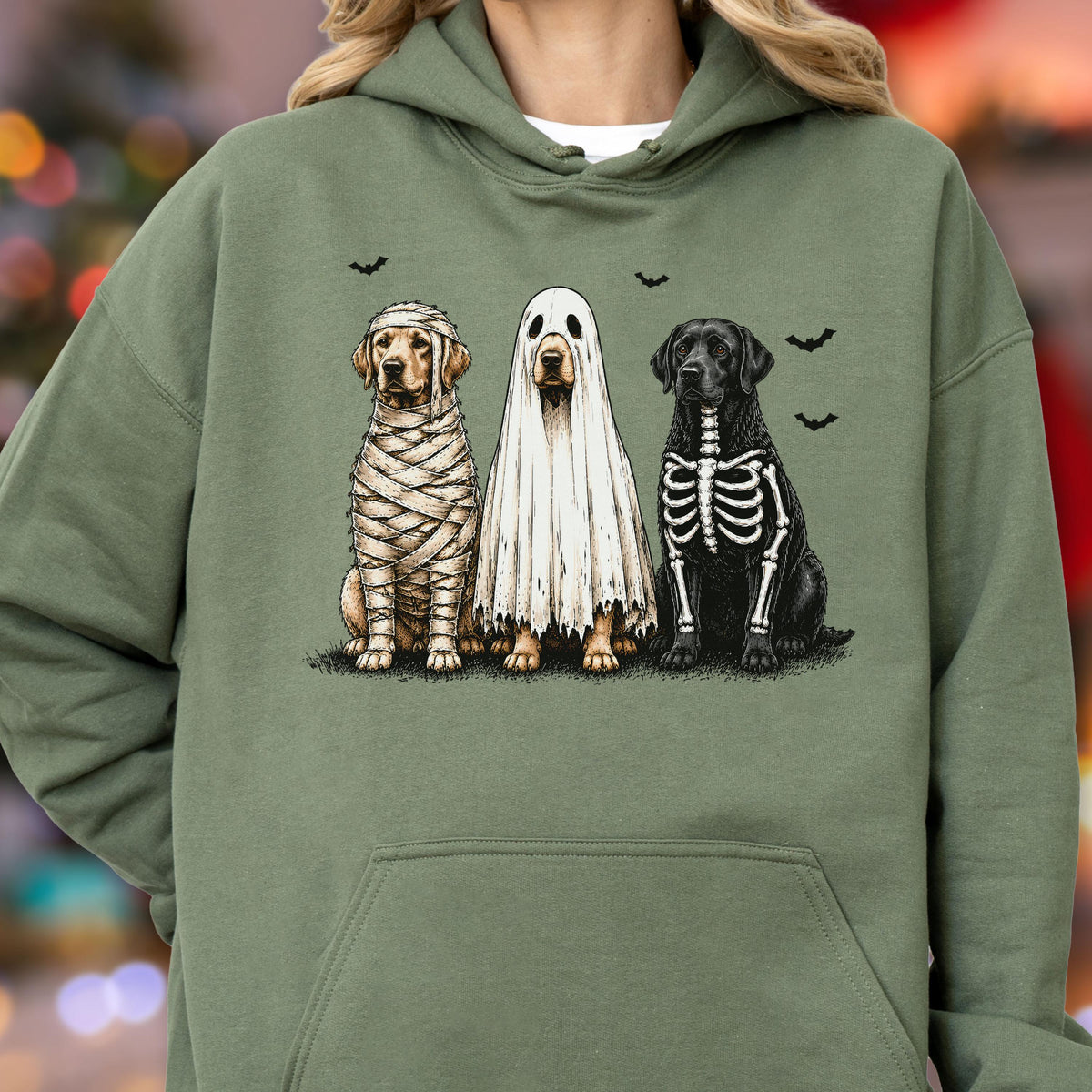 May include: An olive green hooded sweatshirt featuring a graphic print of three dogs in Halloween costumes. One dog is wrapped like a mummy, one is wearing a white ghost sheet, and one has a skeleton design printed on its black fur with small bats.