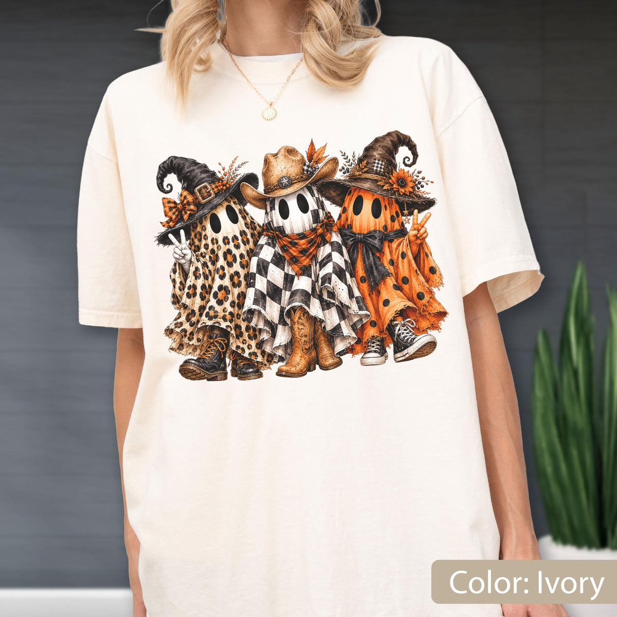 May include: An ivory t-shirt featuring a graphic of three ghosts wearing hats. The left ghost has a leopard print sheet and black boots. The middle ghost has a black and white checkered sheet and cowboy boots. The right ghost has an orange polka dot sheet.