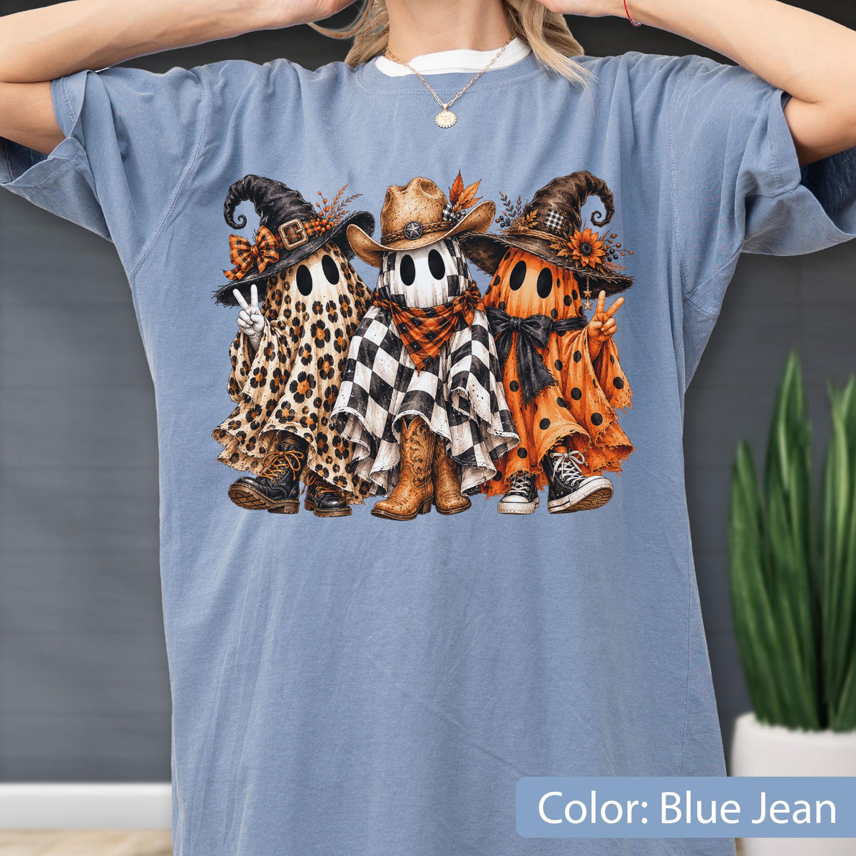 May include: A blue jean colored t-shirt featuring a graphic of three ghosts. The left ghost wears a leopard print sheet, witch hat, and boots. The center ghost wears a black and white checkered sheet, cowboy hat, and boots. The right ghost wears an orange polka dot sheet, witch hat, and sneakers.