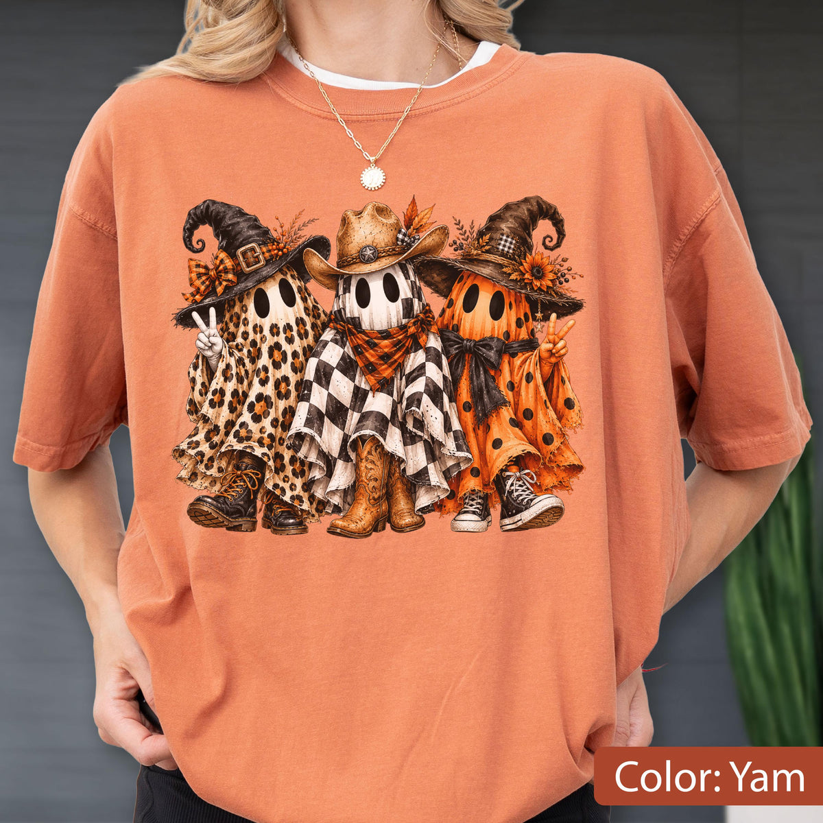 May include: An orange t-shirt featuring a graphic of three ghosts wearing hats and boots. The left ghost has a leopard print sheet, the middle ghost has a black and white checkered sheet with a cowboy hat, and the right ghost has an orange polka dot sheet.
