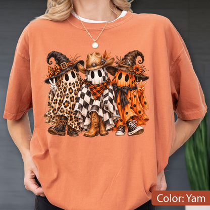 May include: An orange t-shirt featuring a graphic of three ghosts wearing hats and boots. The left ghost has a leopard print sheet, the middle ghost has a black and white checkered sheet with a cowboy hat, and the right ghost has an orange polka dot sheet.