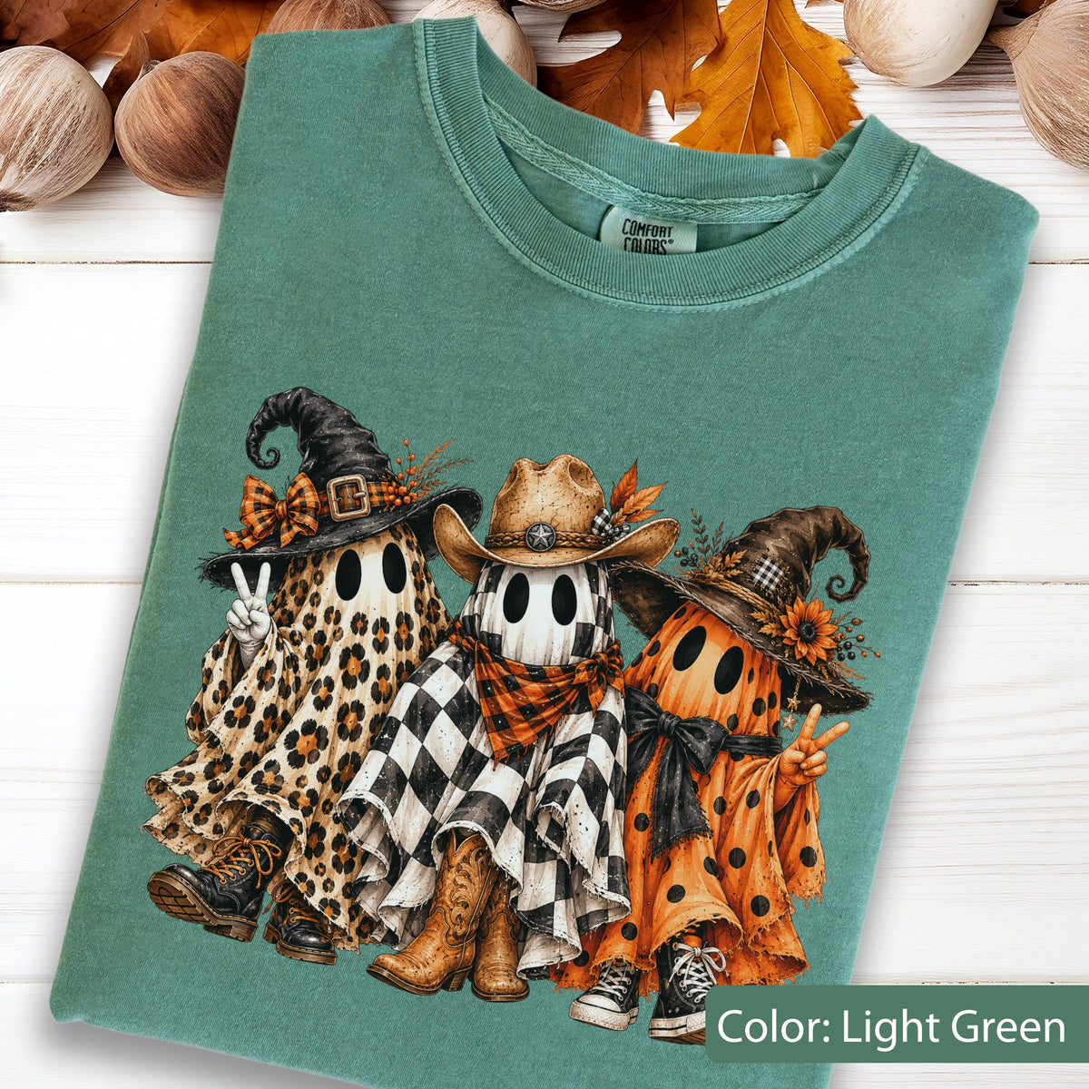May include: A light green t-shirt featuring a graphic of three ghosts. The left ghost wears a leopard print sheet, witch hat, and boots. The center ghost wears a black and white checkered sheet, cowboy hat, and boots. The right ghost wears an orange polka dot sheet, witch hat, and sneakers.