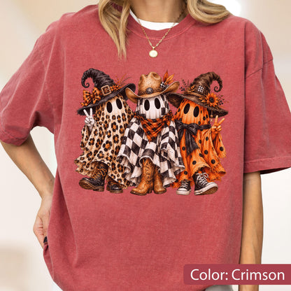 May include: A crimson t-shirt featuring a graphic of three ghosts wearing hats. The left ghost has a leopard print sheet and combat boots, the center ghost has a checkered sheet and cowboy boots, and the right ghost has an orange polka dot sheet.