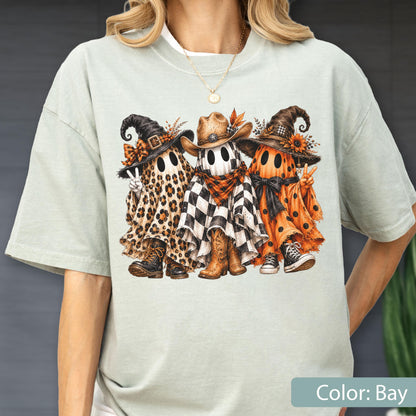 May include: A light sage green t-shirt featuring a graphic of three ghosts wearing hats. The left ghost has a leopard print sheet and boots, the center ghost wears a cowboy hat and checkered sheet, and the right ghost has an orange polka dot sheet.