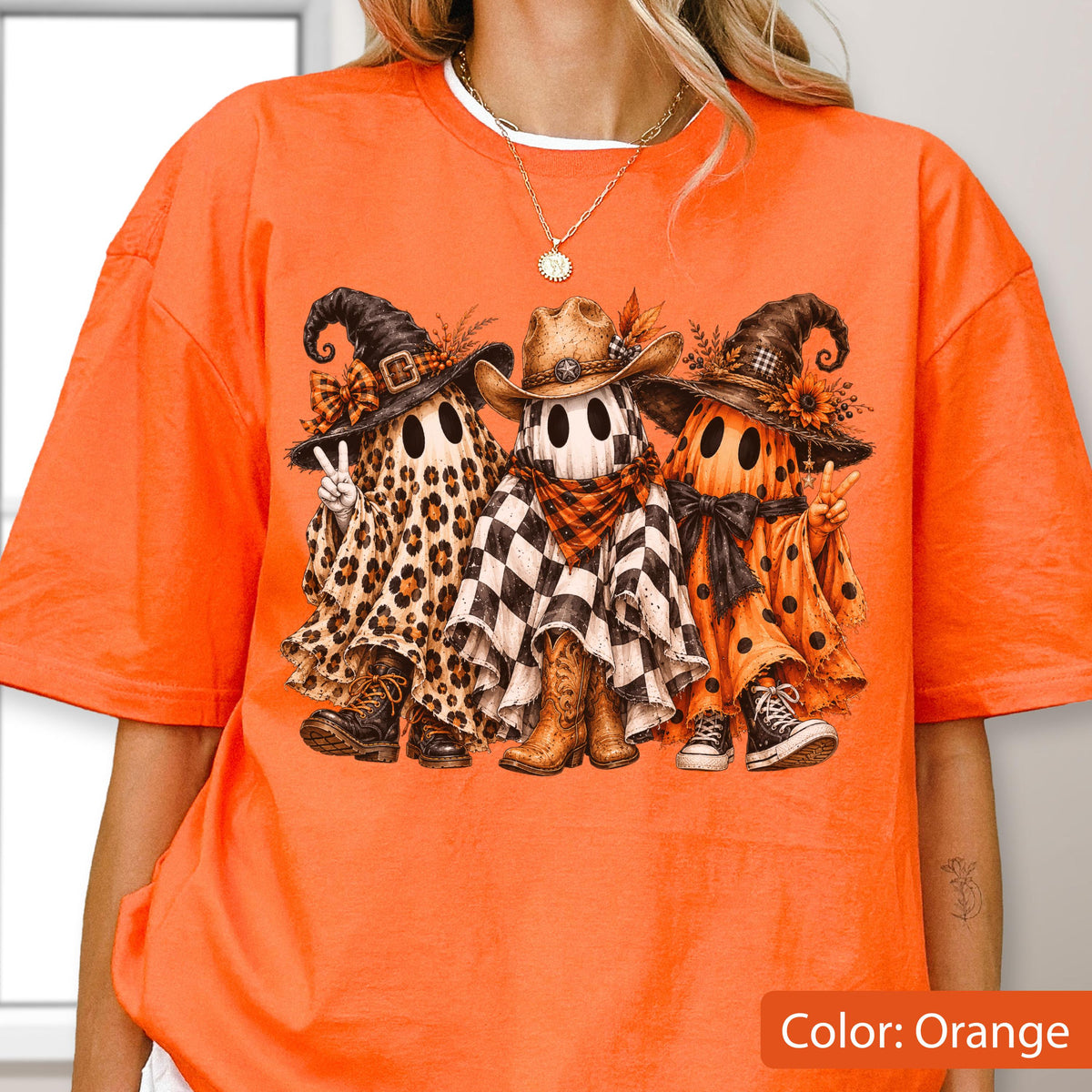 May include: An orange t-shirt featuring a graphic of three ghosts wearing hats and boots. The left ghost has a leopard print sheet, the middle ghost has a black and white checkered sheet with a cowboy hat, and the right ghost has an orange polka dot sheet.